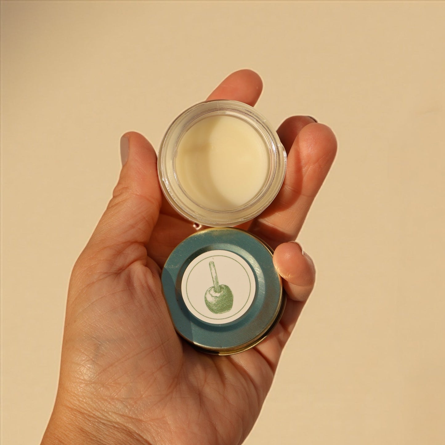 Hand holding a small open container of cream against a beige background